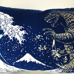 May include: A blue pillow with a white wave design and a Godzilla-like monster in the background. The monster is facing the wave and has a large mouth with sharp teeth. The pillow is decorated with white stars.