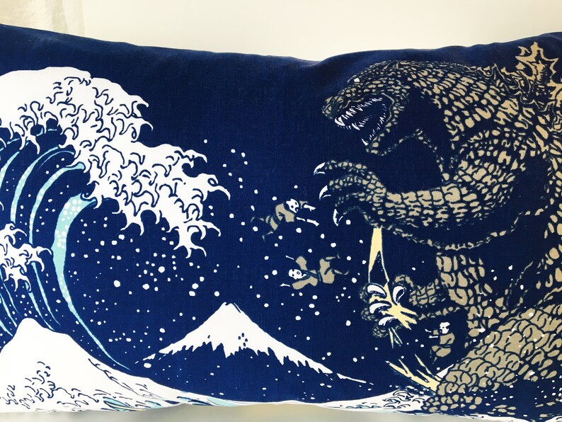 May include: A blue pillow with a white wave design and a Godzilla-like monster in the background. The monster is facing the wave and has a large mouth with sharp teeth. The pillow is decorated with white stars.