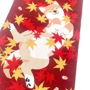 May include: A rectangular fabric runner featuring a deep red background with a printed illustration of a light brown and white Shiba Inu dog lying on its back, surrounded by scattered vibrant red and yellow maple leaves and small brown acorns.