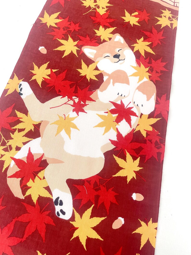 May include: A rectangular fabric runner featuring a deep red background with a printed illustration of a light brown and white Shiba Inu dog lying on its back, surrounded by scattered vibrant red and yellow maple leaves and small brown acorns.