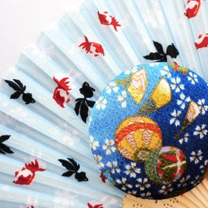 May include: A folding hand fan with a light blue background featuring a pattern of red and black goldfish, partially covered by a circular, fabric-covered button with a dark blue base, white cherry blossom motifs, and colorful spherical patterns.