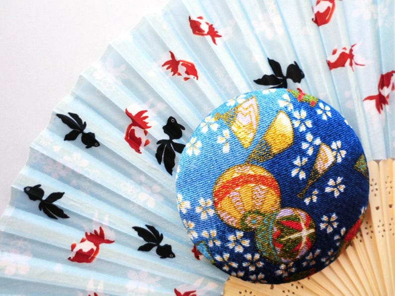 May include: A folding hand fan with a light blue background featuring a pattern of red and black goldfish, partially covered by a circular, fabric-covered button with a dark blue base, white cherry blossom motifs, and colorful spherical patterns.