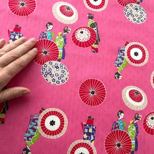 May include: Pink fabric with a repeating pattern of women in traditional Japanese clothing holding umbrellas. The umbrellas are red, white, and black with a floral design. The background is a light pink with a subtle polka dot pattern.