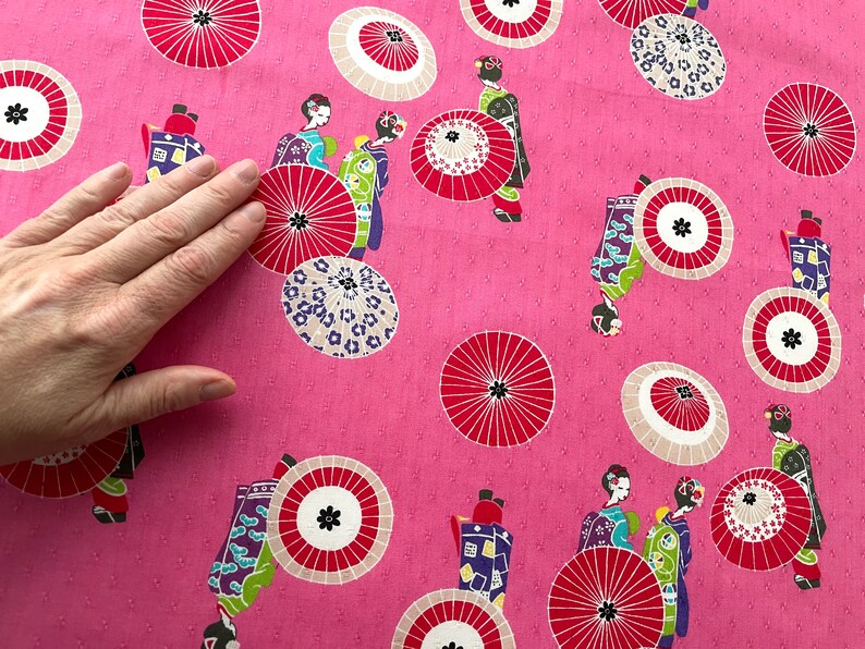 May include: Pink fabric with a repeating pattern of women in traditional Japanese clothing holding umbrellas. The umbrellas are red, white, and black with a floral design. The background is a light pink with a subtle polka dot pattern.