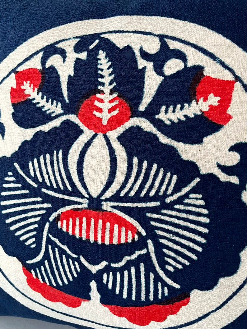 Op de afbeelding: Een blauw en wit kussen met een rood bloemenontwerp. Het kussen heeft een gestileerde bloem met een centrale stengel en bladeren.