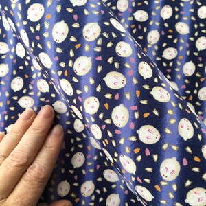 May include: Close-up of a navy blue fabric with a repeating pattern of white bunny faces and scattered floral shapes in pink and orange. The fabric has a slight sheen and is gathered in folds.