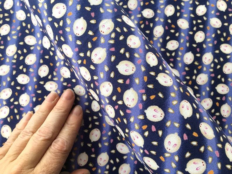 May include: Close-up of a navy blue fabric with a repeating pattern of white bunny faces and scattered floral shapes in pink and orange. The fabric has a slight sheen and is gathered in folds.