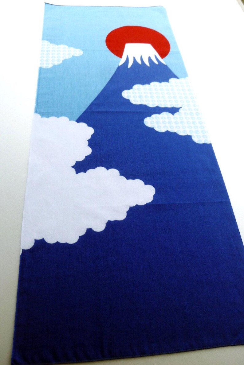 May include: A rectangular textile featuring a graphic print of a mountain with a white snow-capped peak against a large red sun. The background transitions from light blue at the top to dark blue at the bottom, accented by white clouds.