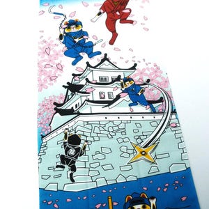 May include: A rectangular fabric panel featuring a colorful illustration of cats dressed as ninjas in action around a white castle. The scene includes falling pink cherry blossoms, a yellow crescent moon, and blue gradient sky and water elements.