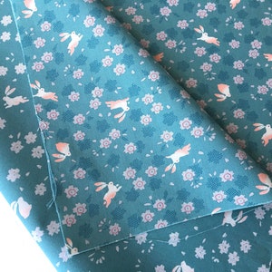 Peut inclure: Tissu bleu canard avec un motif r&eacute;p&eacute;t&eacute; de lapins blancs et de petites fleurs roses. Le tissu est pli&eacute;, r&eacute;v&eacute;lant le motif en d&eacute;tail. Le motif comprend de petits carr&eacute;s bleu-gris.