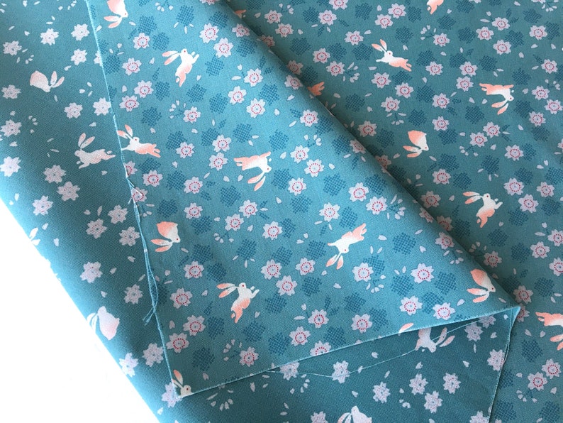 Peut inclure: Tissu bleu canard avec un motif r&eacute;p&eacute;t&eacute; de lapins blancs et de petites fleurs roses. Le tissu est pli&eacute;, r&eacute;v&eacute;lant le motif en d&eacute;tail. Le motif comprend de petits carr&eacute;s bleu-gris.