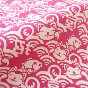 Pu&ograve; includere: Un tessuto rosa vivace con un motivo bianco ripetuto di gatti portafortuna, pesci stilizzati e onde vorticose. Il design include caratteri kanji giapponesi integrati nella fitta stampa grafica che copre l'intera superficie del tessuto.