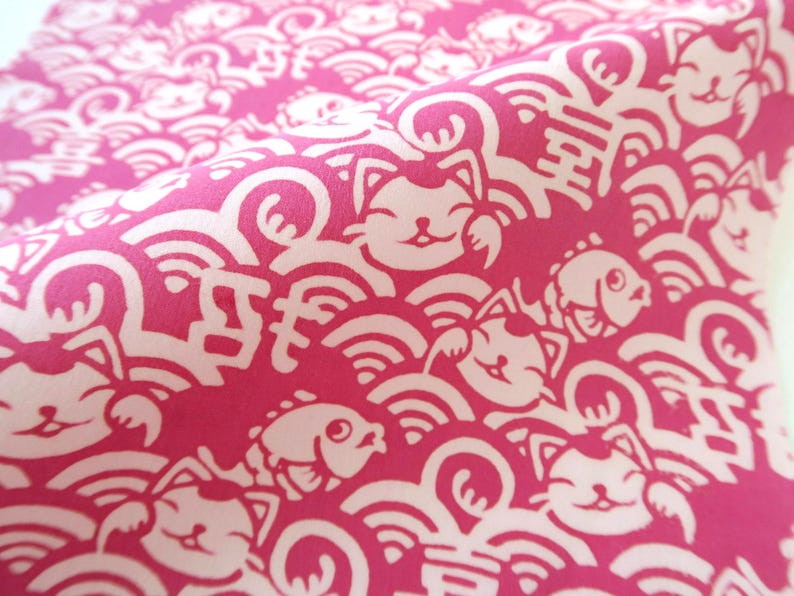 Pu&ograve; includere: Un tessuto rosa vivace con un motivo bianco ripetuto di gatti portafortuna, pesci stilizzati e onde vorticose. Il design include caratteri kanji giapponesi integrati nella fitta stampa grafica che copre l'intera superficie del tessuto.