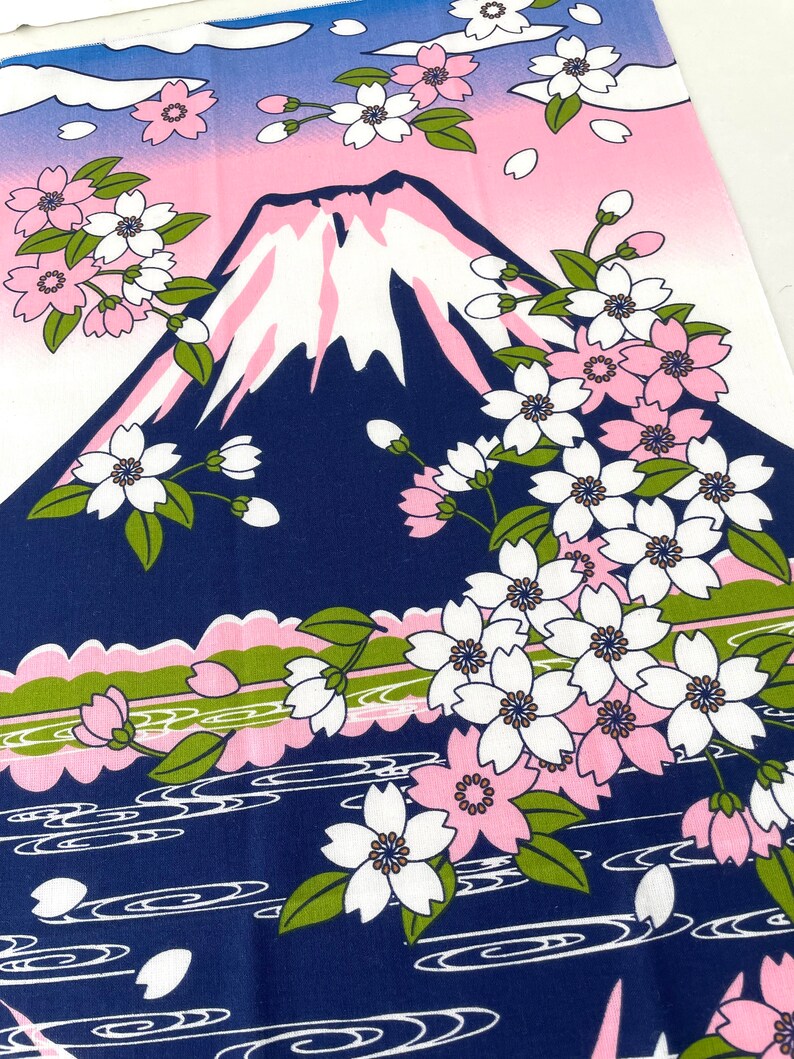 May include: A rectangular fabric textile featuring a graphic illustration of a dark blue mountain with a white snow-capped peak. The scene includes clusters of white and pink cherry blossoms with green leaves, set against a pink and blue sky.