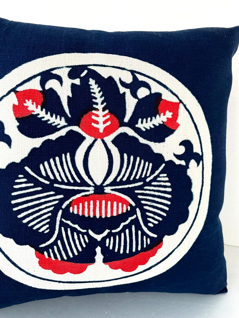 Op de afbeelding: Een marineblauw kussen met een wit cirkelvormig ontwerp met een gestileerd rood en zwart bloemenpatroon.
