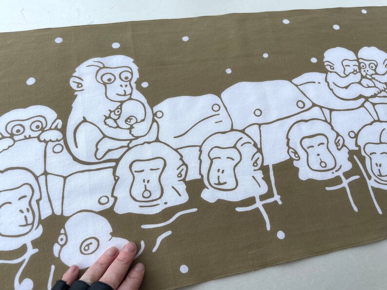 May include: A brown and white fabric with a repeating pattern of cartoon monkeys. The monkeys are white with brown outlines and are depicted in various poses, some holding babies. The background is a solid brown color with white dots.