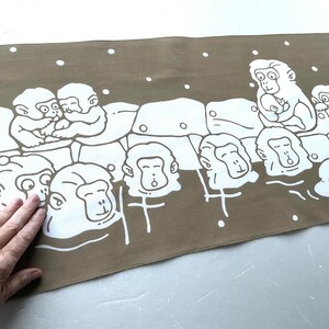 May include: A rectangular, taupe-colored fabric with a white illustration of monkeys in a hot spring. The monkeys are depicted in various poses, some cuddling, others with their heads above the water. White dots are scattered across the top.