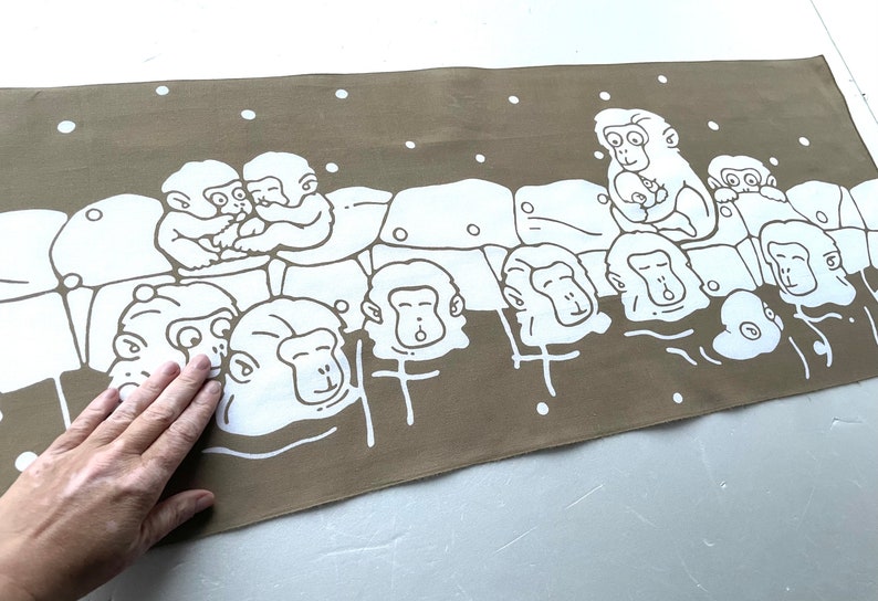 May include: A rectangular, taupe-colored fabric with a white illustration of monkeys in a hot spring. The monkeys are depicted in various poses, some cuddling, others with their heads above the water. White dots are scattered across the top.
