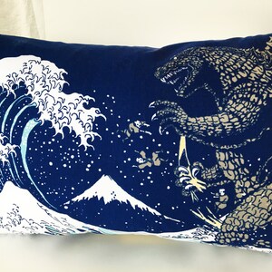 May include: A blue pillow with a white wave and a Godzilla-like monster printed on it. The wave is reminiscent of the Great Wave off Kanagawa, a famous Japanese woodblock print.