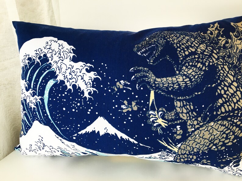 May include: A blue pillow with a white wave and a Godzilla-like monster printed on it. The wave is reminiscent of the Great Wave off Kanagawa, a famous Japanese woodblock print.