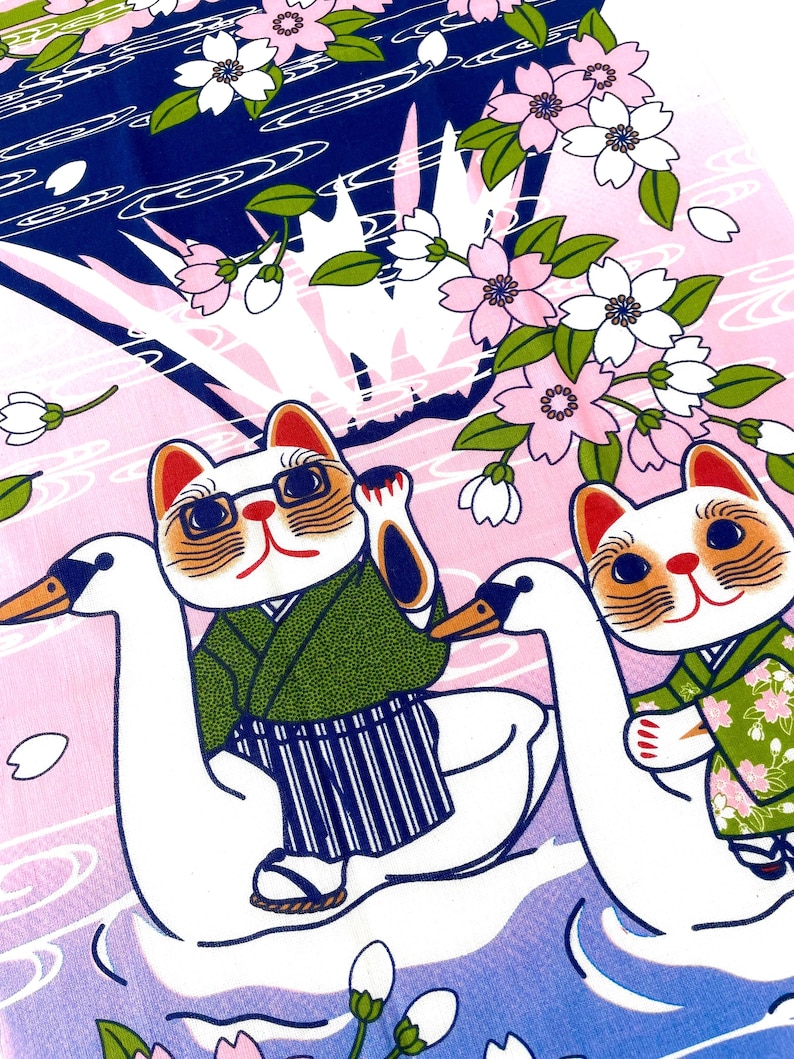 May include: A fabric print featuring two white cats wearing green kimonos, each riding a white swan. The cats are surrounded by pink cherry blossoms and white petals against a background of dark blue and light pink water ripples.