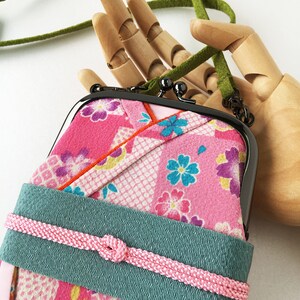 May include: A rectangular pouch with a black metal kiss-lock frame, featuring a pink fabric exterior with a multi-colored floral pattern. A wide teal band wraps around the center, secured by a thin pink braided cord. It has a green strap.