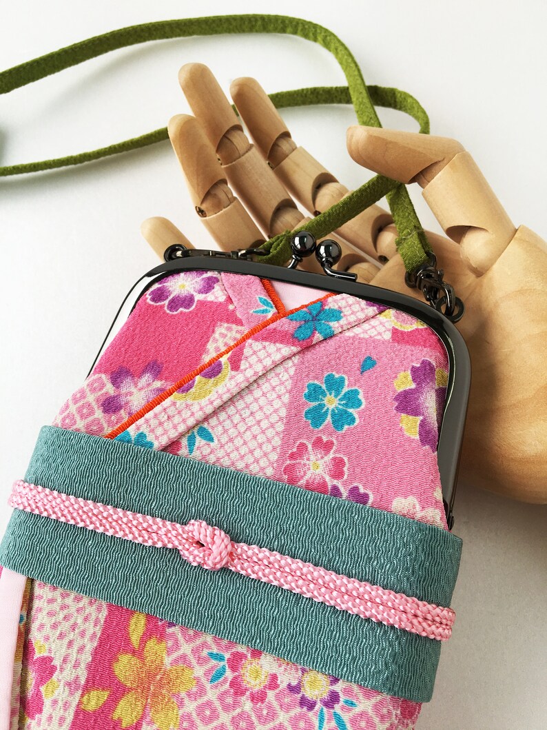 May include: A rectangular pouch with a black metal kiss-lock frame, featuring a pink fabric exterior with a multi-colored floral pattern. A wide teal band wraps around the center, secured by a thin pink braided cord. It has a green strap.