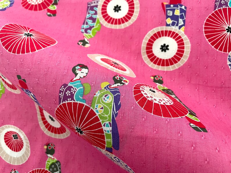 May include: Pink fabric with a repeating pattern of geishas and red and white umbrellas. The geishas are wearing traditional Japanese clothing and carrying umbrellas.
