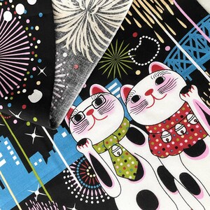 May include: Two white lucky cat figurines with black spots and pink cheeks, wearing a green and black polka dot tie and a red and white polka dot dress, against a black background with blue and white city skyline and fireworks.