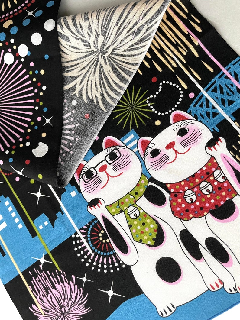 May include: Two white lucky cat figurines with black spots and pink cheeks, wearing a green and black polka dot tie and a red and white polka dot dress, against a black background with blue and white city skyline and fireworks.