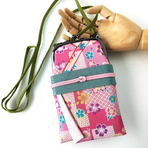 May include: A rectangular fabric pouch with a metal frame and clasp, featuring a pink floral pattern with geometric accents. It includes a wide teal band across the center, a thin pink cord tie, and a long green strap for carrying over the shoulder.