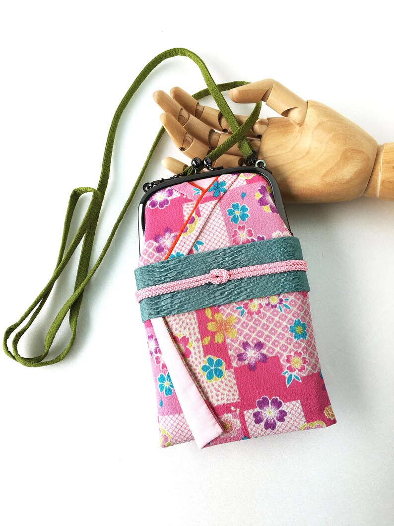 May include: A rectangular fabric pouch with a metal frame and clasp, featuring a pink floral pattern with geometric accents. It includes a wide teal band across the center, a thin pink cord tie, and a long green strap for carrying over the shoulder.