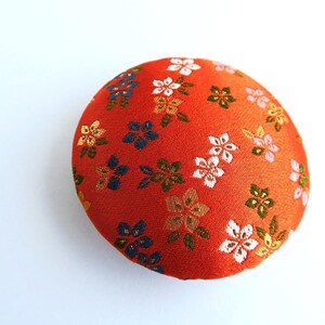 May include: A round, orange fabric pincushion featuring a repeating pattern of small, embroidered flowers in shades of white, blue, green, and gold. The surface has a textured, woven appearance with a dense arrangement of floral motifs.