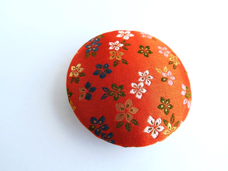 May include: A round, orange fabric pincushion featuring a repeating pattern of small, embroidered flowers in shades of white, blue, green, and gold. The surface has a textured, woven appearance with a dense arrangement of floral motifs.