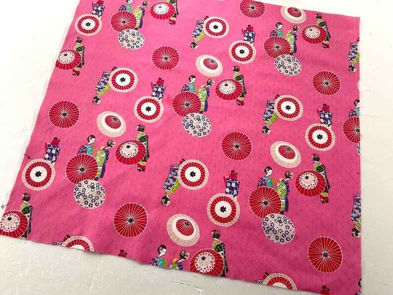 May include: Pink fabric with a repeating pattern of geishas and umbrellas. The geishas are wearing colorful kimonos and holding parasols. The umbrellas are red, white, and black.