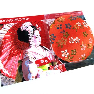 May include: A round fabric brooch featuring a vibrant orange background with a repeating pattern of small white, green, and blue floral motifs. The brooch is presented in clear packaging alongside a card labeled KIMONO BROOCH with an image of a person.