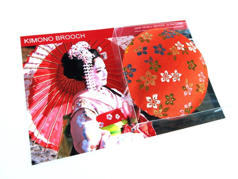 May include: A round fabric brooch featuring a vibrant orange background with a repeating pattern of small white, green, and blue floral motifs. The brooch is presented in clear packaging alongside a card labeled KIMONO BROOCH with an image of a person.