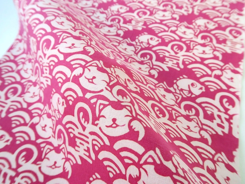 Pu&ograve; includere: Un tessuto rosa vivace con un motivo ripetuto di gatti portafortuna bianchi, pesci stilizzati e onde. Il design include piccoli caratteri kanji intervallati tra i gatti e i pesci, creando una stampa grafica densa e continua.