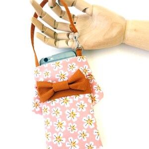 May include: A rectangular phone pouch featuring a light pink fabric with a repeating white and yellow flower pattern. The pouch includes a solid brown bow accent on the front and a matching brown wrist strap attached with a silver metal swivel clasp.