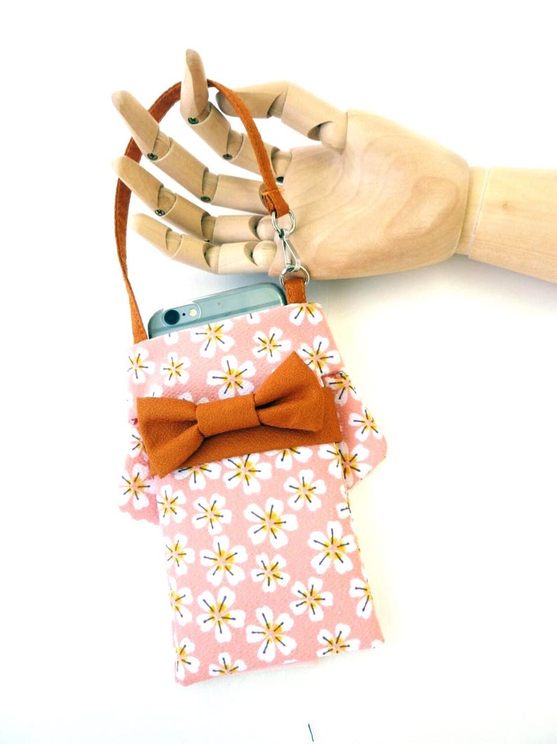 May include: A rectangular phone pouch featuring a light pink fabric with a repeating white and yellow flower pattern. The pouch includes a solid brown bow accent on the front and a matching brown wrist strap attached with a silver metal swivel clasp.