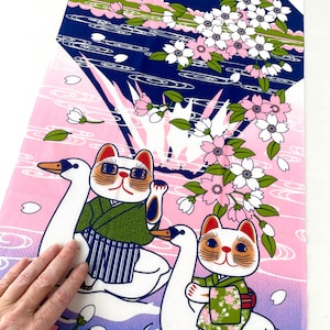 May include: A rectangular fabric panel featuring a graphic print of two cats wearing green patterned kimonos, each riding a white swan. The background displays a dark blue mountain silhouette, pink gradient water, and white cherry blossom branches.