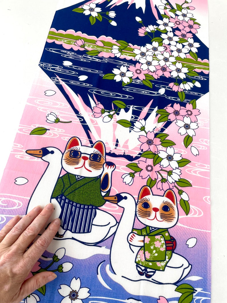 May include: A rectangular fabric panel featuring a graphic print of two cats wearing green patterned kimonos, each riding a white swan. The background displays a dark blue mountain silhouette, pink gradient water, and white cherry blossom branches.