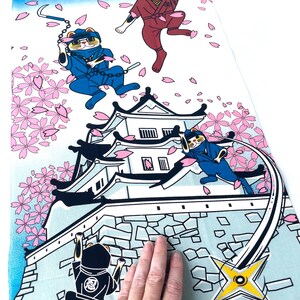 May include: A rectangular fabric panel featuring a colorful illustration of cats dressed as ninjas in blue and red outfits. The scene includes a white castle, falling pink cherry blossoms, a yellow crescent moon, and shuriken stars.
