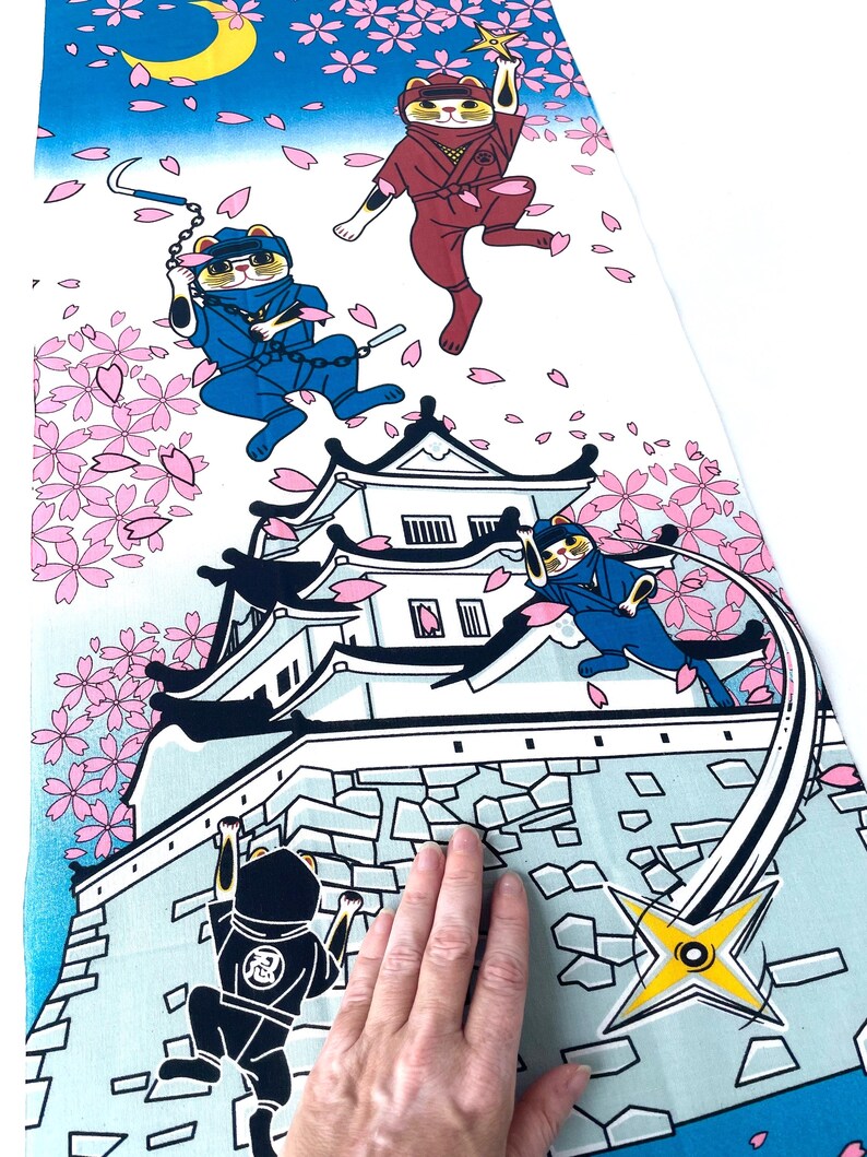 May include: A rectangular fabric panel featuring a colorful illustration of cats dressed as ninjas in blue and red outfits. The scene includes a white castle, falling pink cherry blossoms, a yellow crescent moon, and shuriken stars.