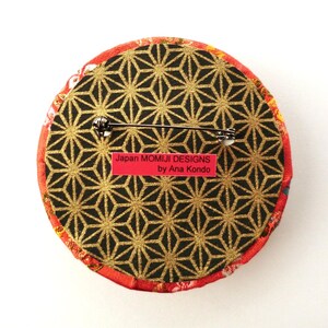 May include: A circular brooch featuring a black and gold geometric star pattern on the back, with a metal pin mechanism. A rectangular red label is attached, displaying the text Japan MOMIJI DESIGNS by Ana Kondo in black lettering.