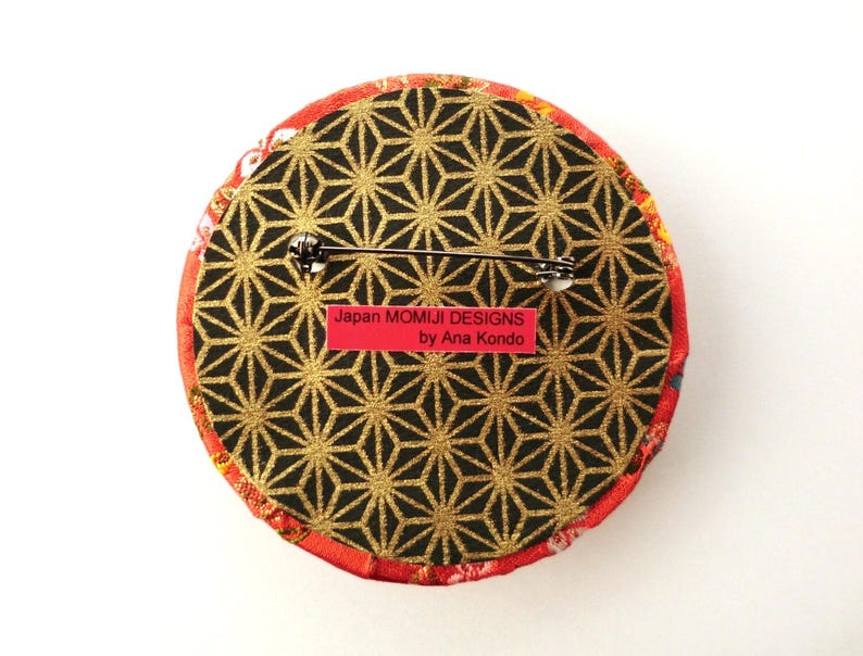 May include: A circular brooch featuring a black and gold geometric star pattern on the back, with a metal pin mechanism. A rectangular red label is attached, displaying the text Japan MOMIJI DESIGNS by Ana Kondo in black lettering.