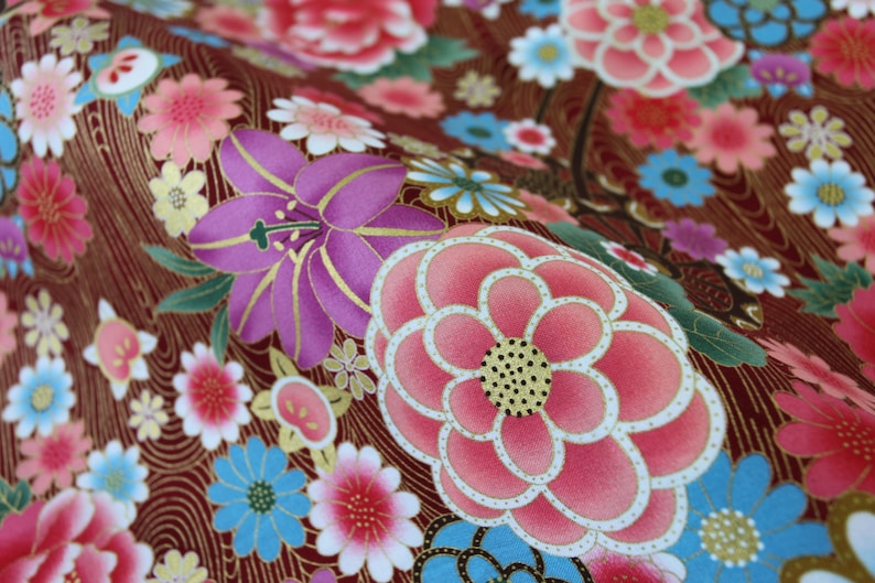 May include: A fabric featuring a deep maroon background with a gold-toned wavy line pattern. The surface is covered in a dense floral print with large pink and purple blossoms, smaller light blue and white flowers, and green leaves with gold outlines.