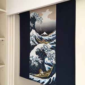 May include: A blue and white tapestry featuring a large wave with a boat and a small figure in the foreground. The wave is reminiscent of the Great Wave off Kanagawa by Hokusai. The tapestry is hanging on a white wall.