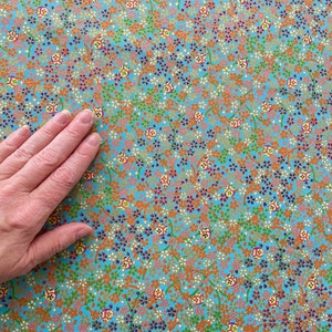 Puede incluir: Una tela de color azul claro con un patr&oacute;n denso y repetitivo de peque&ntilde;as flores naranjas, verdes y moradas oscuras, junto con diminutos motivos circulares que contienen dise&ntilde;os geom&eacute;tricos amarillos, rojos y blancos.