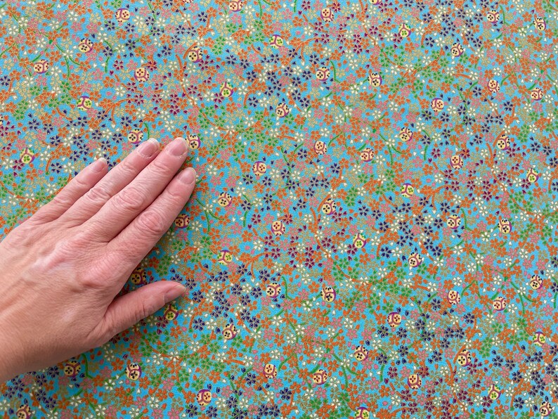 Puede incluir: Una tela de color azul claro con un patr&oacute;n denso y repetitivo de peque&ntilde;as flores naranjas, verdes y moradas oscuras, junto con diminutos motivos circulares que contienen dise&ntilde;os geom&eacute;tricos amarillos, rojos y blancos.